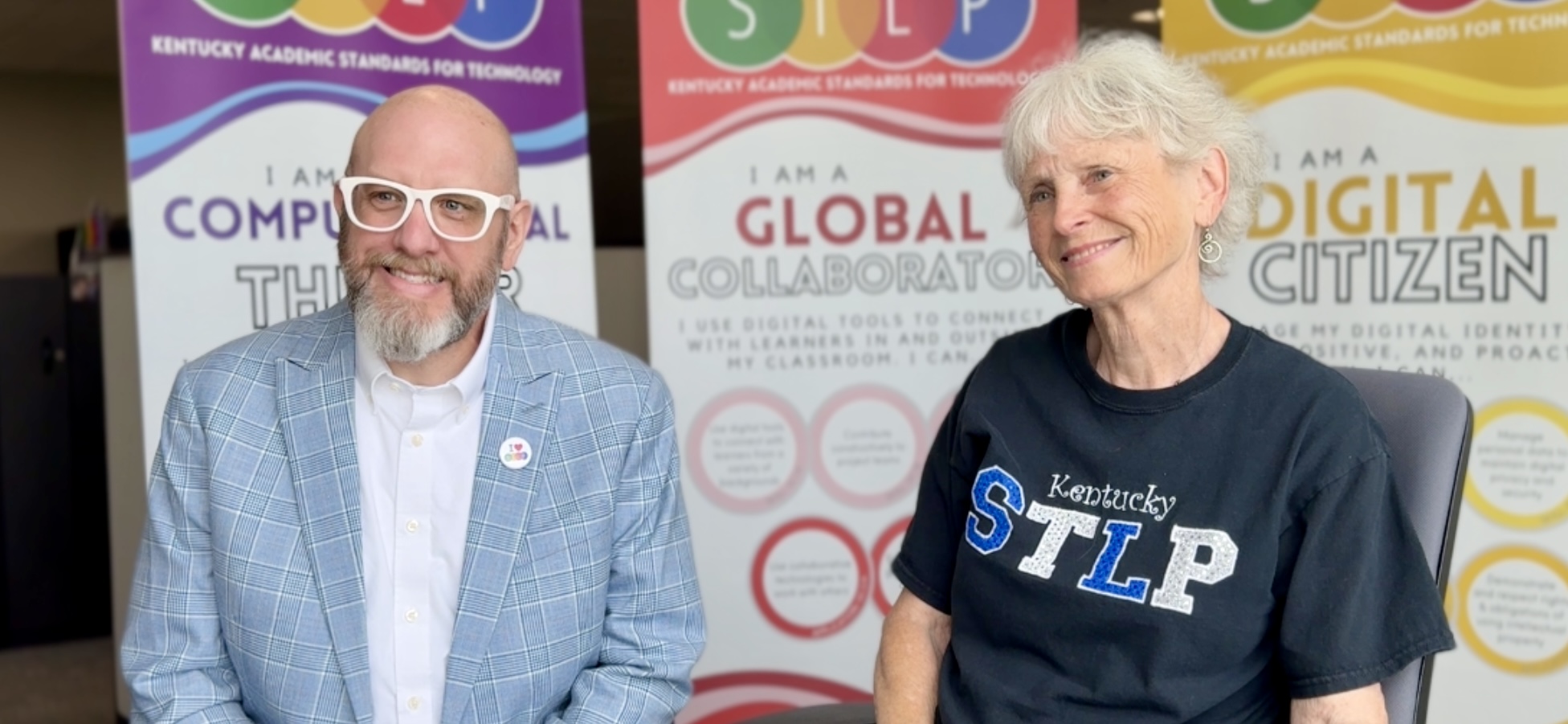 Jeff Sebulsky and Elaine Harrison-Lane, two key figures of Kentucky’s Student Technology Leadership Program (STLP), are seated together smiling during an interview. Jeff is wearing a light blue plaid jacket and glasses, while Elaine is dressed in a Kentucky STLP t-shirt. Behind them are colorful banners promoting Kentucky Academic Standards for Technology, featuring messages like 'Global Collaborator' and 'Digital Citizen.