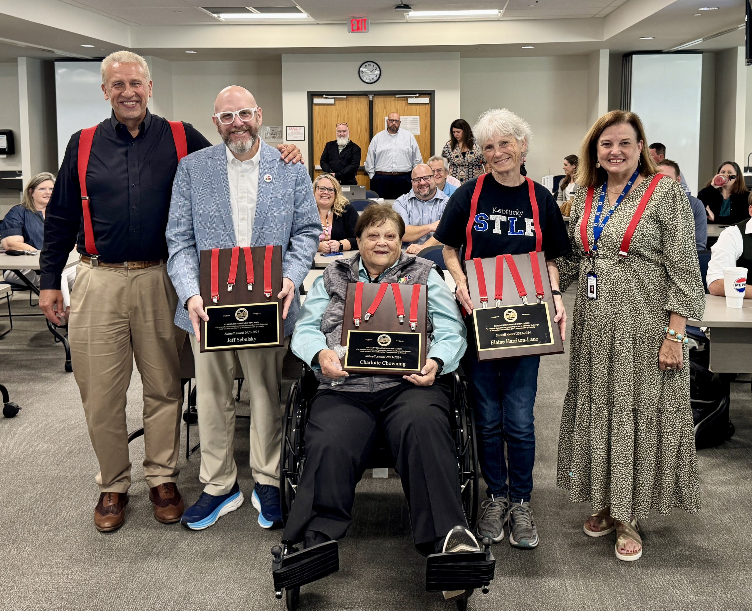 Group photo of the 2023-24 Stilwell Award winners, featuring five individuals holding plaques. The group includes leaders and supporters of Kentucky's Student Technology Leadership Program (STLP), smiling together in a classroom setting. The individuals at the center are honored for their contributions to the program over the years, and they are wearing distinctive red suspenders, symbolizing the award.
